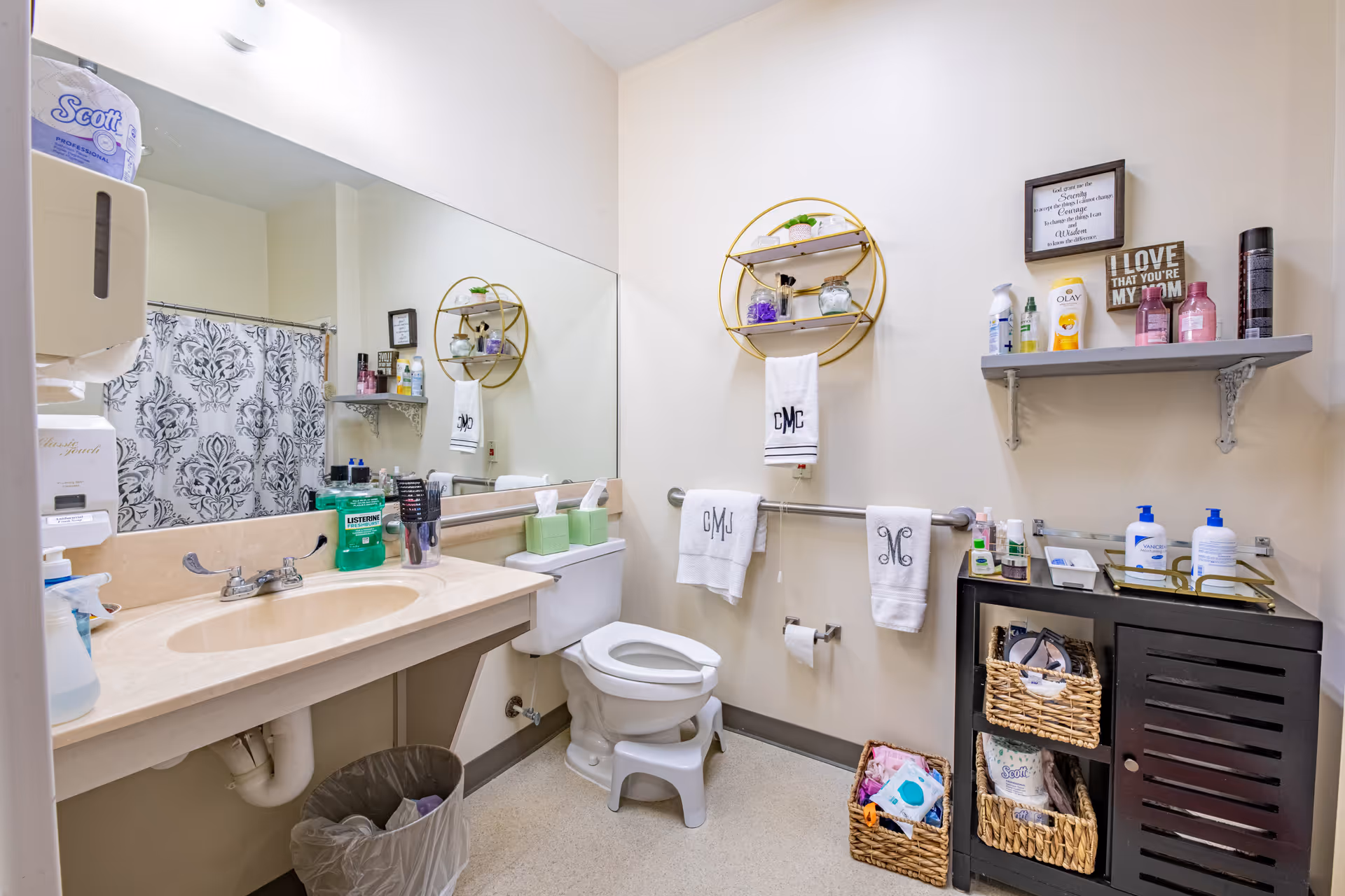 A clean and organized bathroom with a beige countertop sink, a large mirror, and a toilet with a step stool. The walls have a round gold shelf holding toiletries and a towel rack with three white towels. There is a small cabinet with wicker baskets containing various items, and a shelf above it with personal care products and decorative signs. A shower curtain with a black and white pattern is visible in the background.