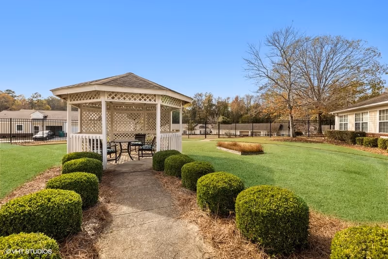 Outdoor garden area with a white wooden gazebo featuring lattice sides and a shingled roof. Inside the gazebo are a table and chairs. The gazebo is surrounded by neatly trimmed bushes and green grass, with trees and buildings visible in the background under a clear blue sky.