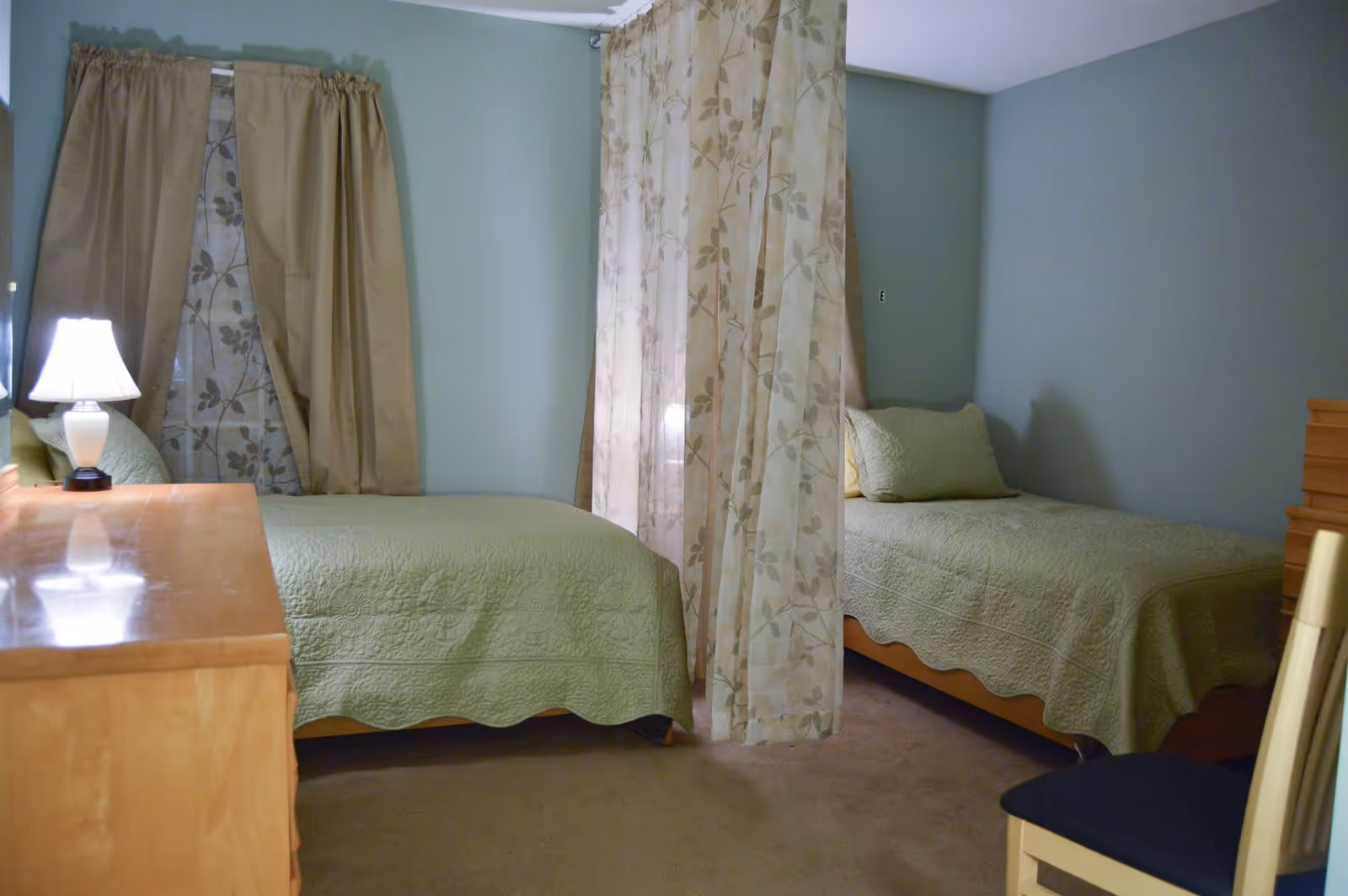 A bedroom with two twin beds separated by a sheer floral curtain. Each bed has a light green quilt and matching pillow. There is a wooden dresser with a lamp on it to the left and a chair with a dark cushion to the right. The walls are painted light green and the window has beige curtains with a floral sheer underneath.