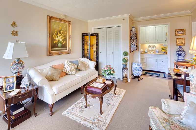 A cozy living room with a white sofa adorned with multiple decorative pillows, a wooden coffee table with books and a flower arrangement, a side table with a lamp and framed photos, floral armchairs, and various decorative items including a large framed painting of flowers, a folding screen, and blue and white ceramic vases. The room has beige walls and carpet, with a small hallway leading to a white cabinet area.
