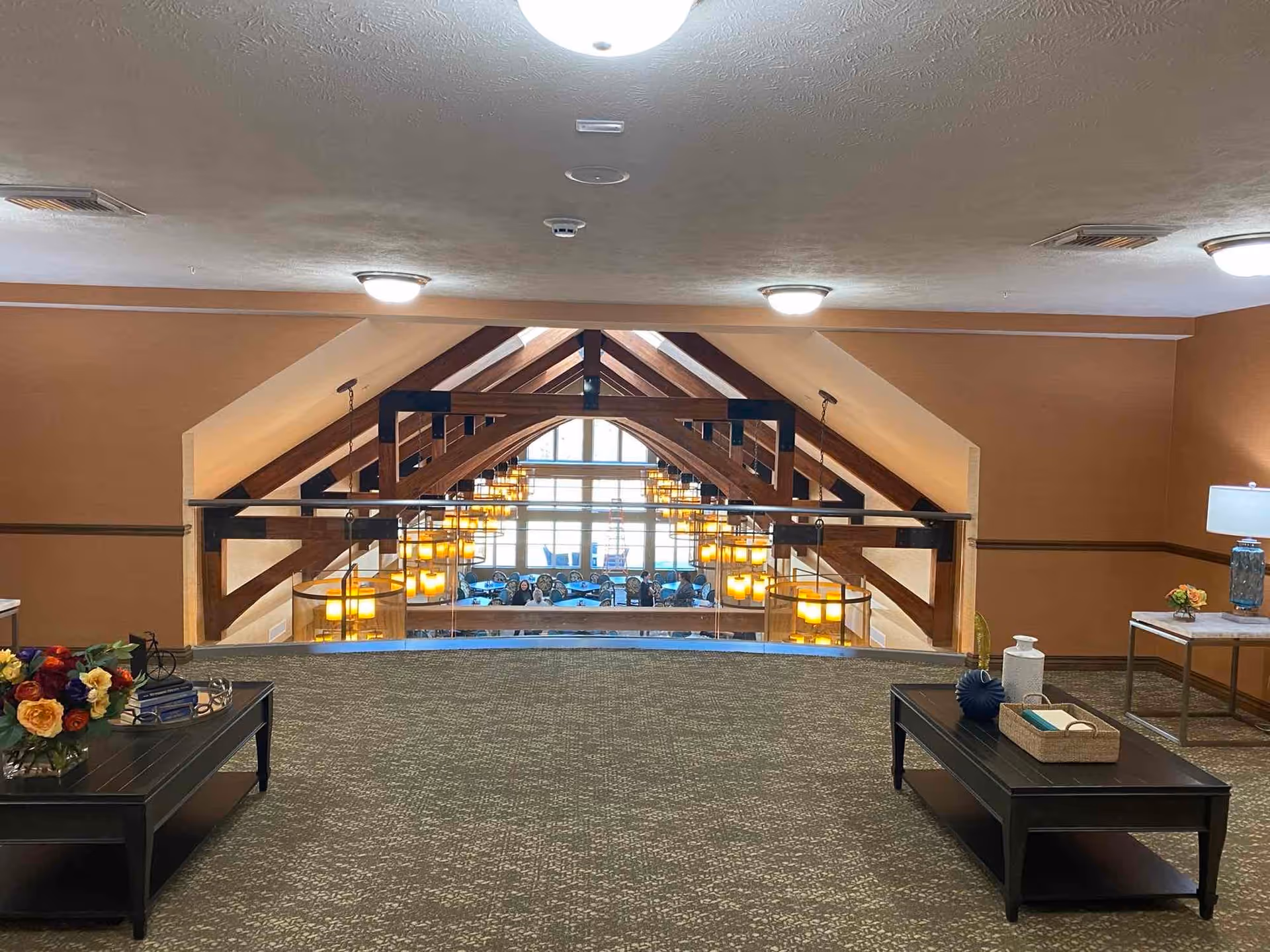 Interior view of a senior living facility showing a carpeted upper hallway with two black coffee tables decorated with flowers, books, and decorative items. The hallway overlooks a large, open dining area below with wooden beams and multiple hanging pendant lights. Large windows in the background let in natural light.