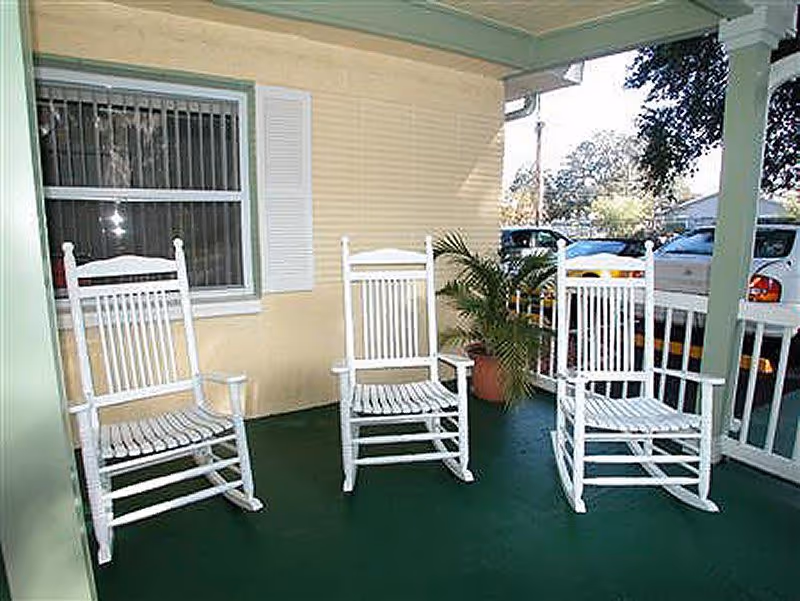 Covered porch with three white rocking chairs on a green floor, a potted plant, and a view of parked cars.