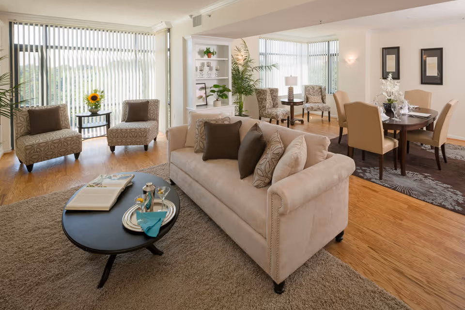 A bright and spacious living room area with a beige sofa adorned with patterned and solid pillows, a black oval coffee table with a book and a tray holding a cocktail and a napkin, two patterned armchairs with brown cushions near large windows with vertical blinds, and a dining area with a round table and six beige chairs on a dark rug. The room features wooden flooring, a built-in white shelving unit with decorative items, and several plants.