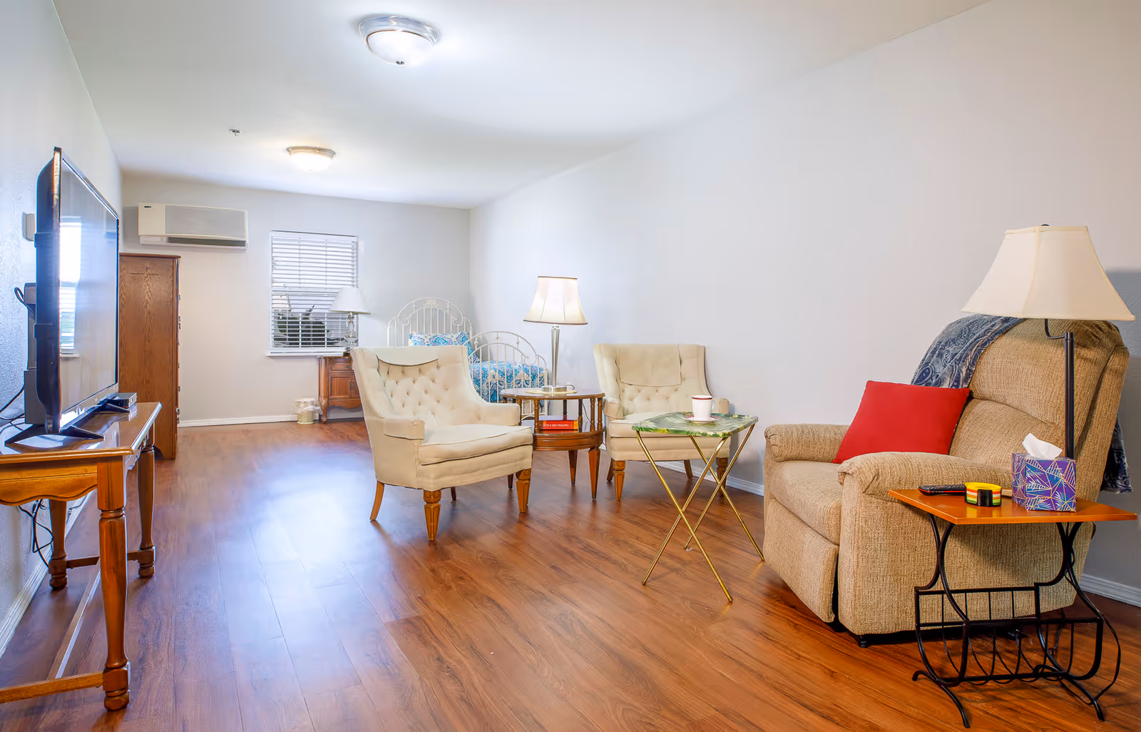 A cozy living area with wooden flooring featuring a beige recliner with a red pillow and a side table with a lamp, tissue box, and remote control. Two cream-colored armchairs and a small round table are positioned in the middle of the room. In the background, there is a bed with a white metal frame, a wooden dresser, a window with blinds, and a wall-mounted air conditioning unit. A flat-screen TV is placed on a wooden table on the left side of the image.