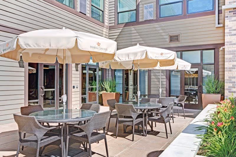 Outdoor patio area at Merrill Gardens at Lafayette with round glass tables, wicker chairs, and large beige umbrellas. The patio is surrounded by the building's exterior walls with large windows and potted plants along the edges.