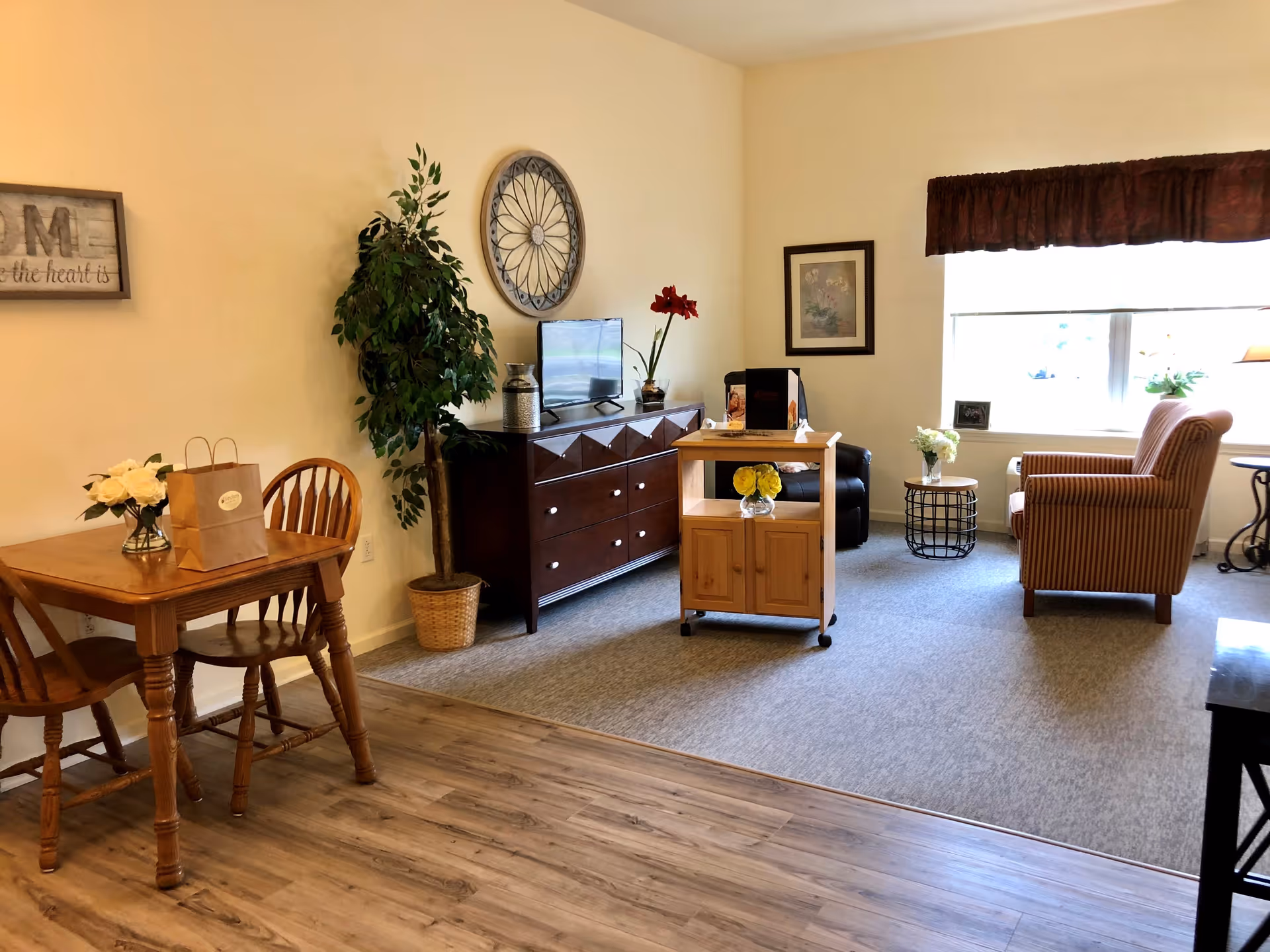 A cozy living room area with a wooden dining table and two chairs on the left, a potted plant next to a dark wooden dresser with a TV on top, a small wooden rolling cart with flowers, a striped armchair near a window with a brown valance, and a small round side table with flowers. The floor transitions from wood to carpet.