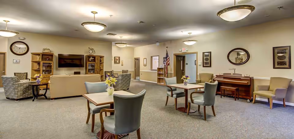 A spacious common area in a senior living facility featuring several tables with chairs and small flower vases, comfortable armchairs, a beige sofa, a wall-mounted flat-screen TV, wooden shelving units, a piano with a bench, and framed pictures on the walls. The room is well-lit with ceiling lights and has a neutral color scheme.