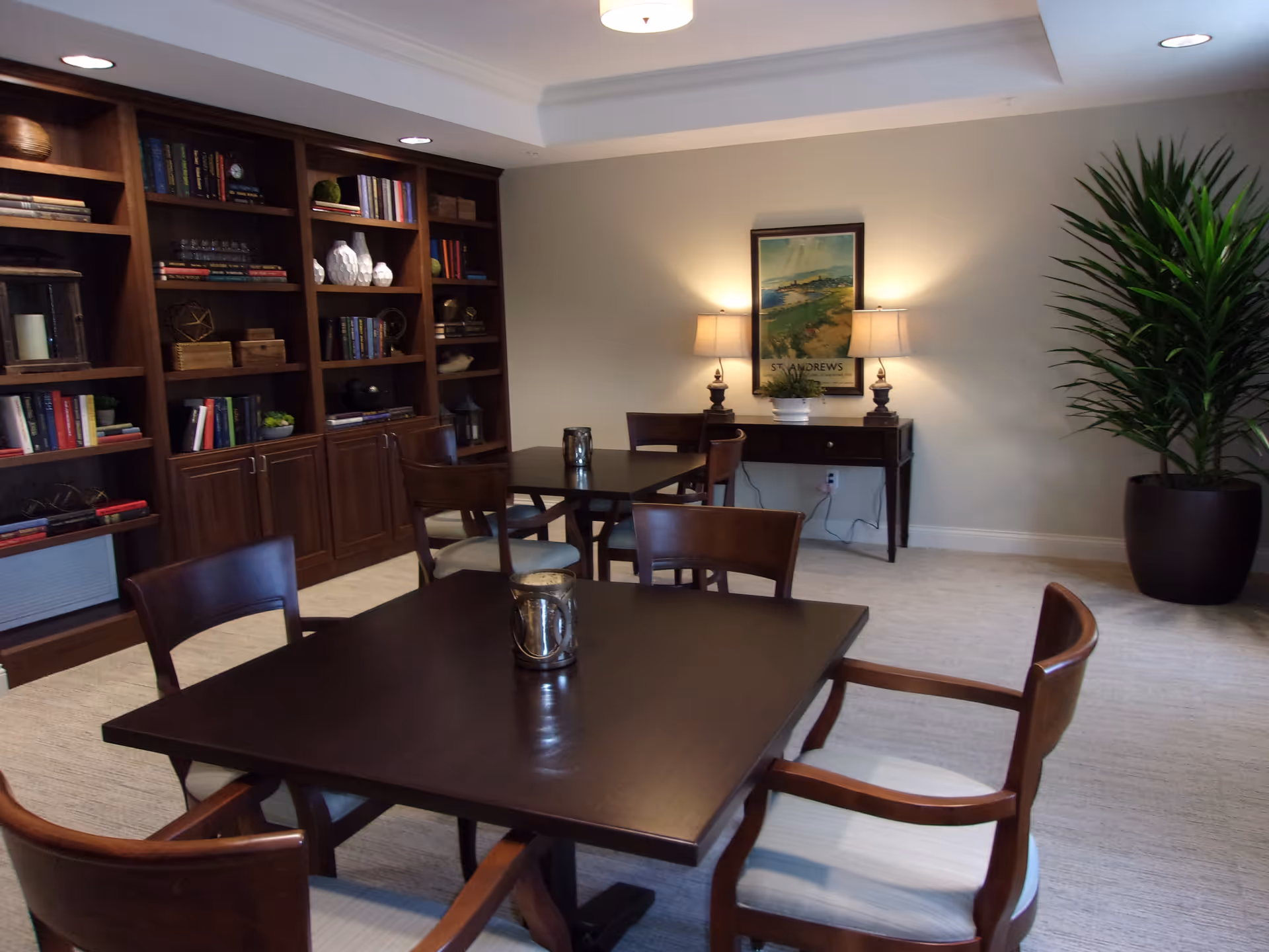 A communal dining/lounge area with dark wood tables and chairs, built-in bookshelves, lamps, and a large potted plant.