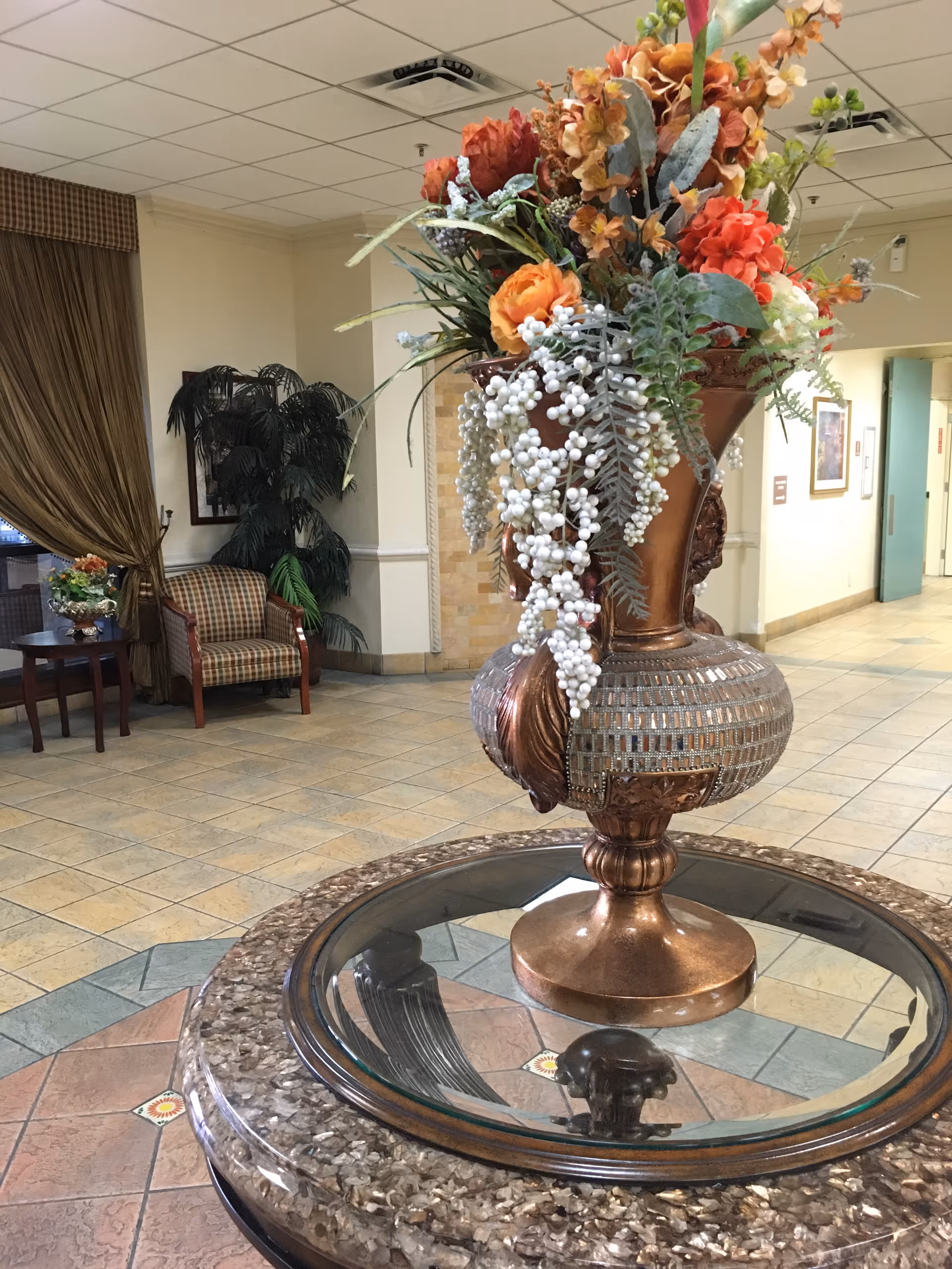 A decorative lobby area featuring a large ornate vase with artificial flowers on a round glass-topped table. In the background, there is a seating area with a plaid upholstered chair, a small table with another floral arrangement, a large potted plant, and brown curtains covering a window. The floor is tiled and the walls are light-colored with some framed artwork.