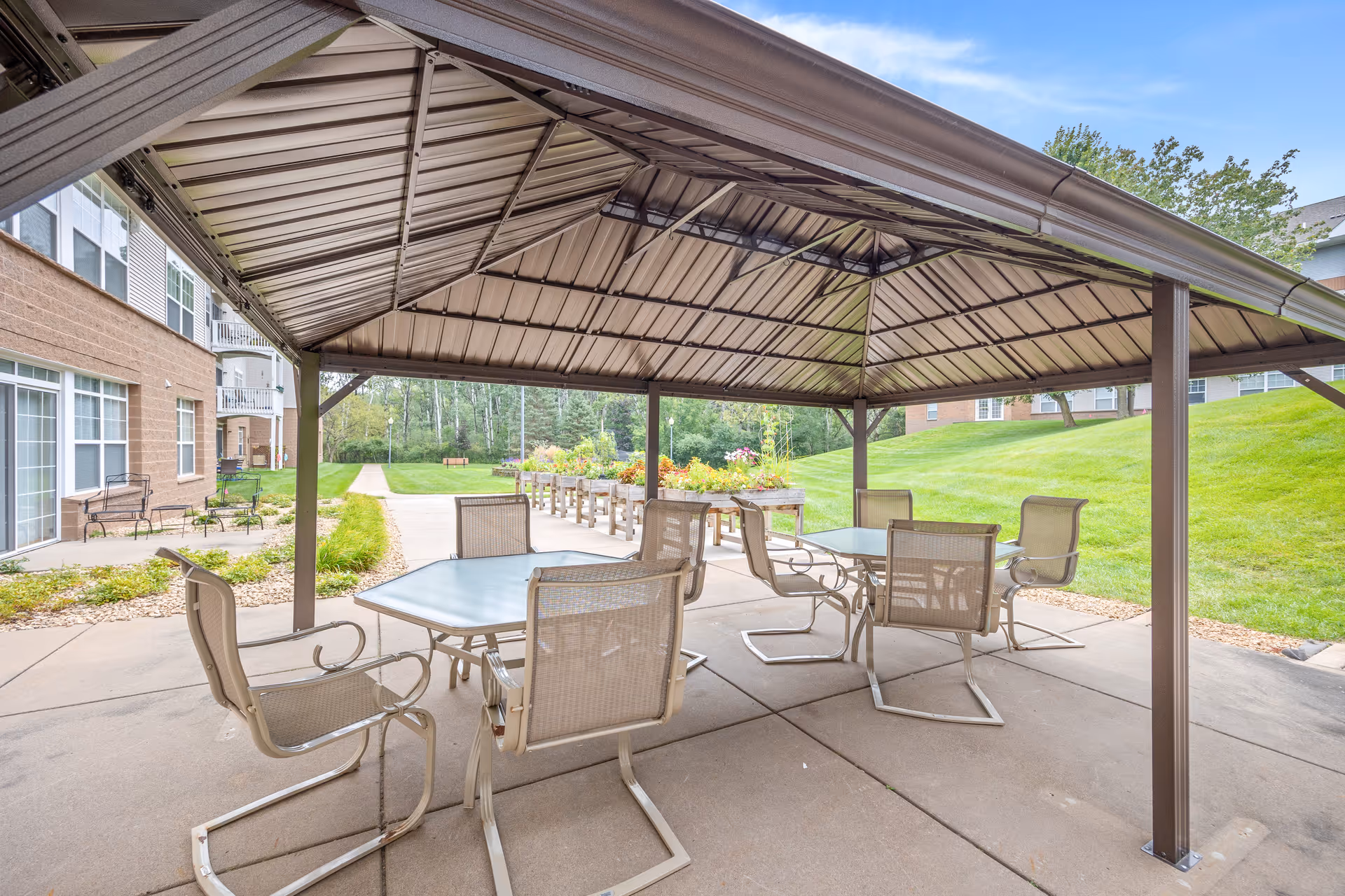 Covered outdoor patio with a metal pavilion sheltering tables and chairs next to a lawn and senior living building.