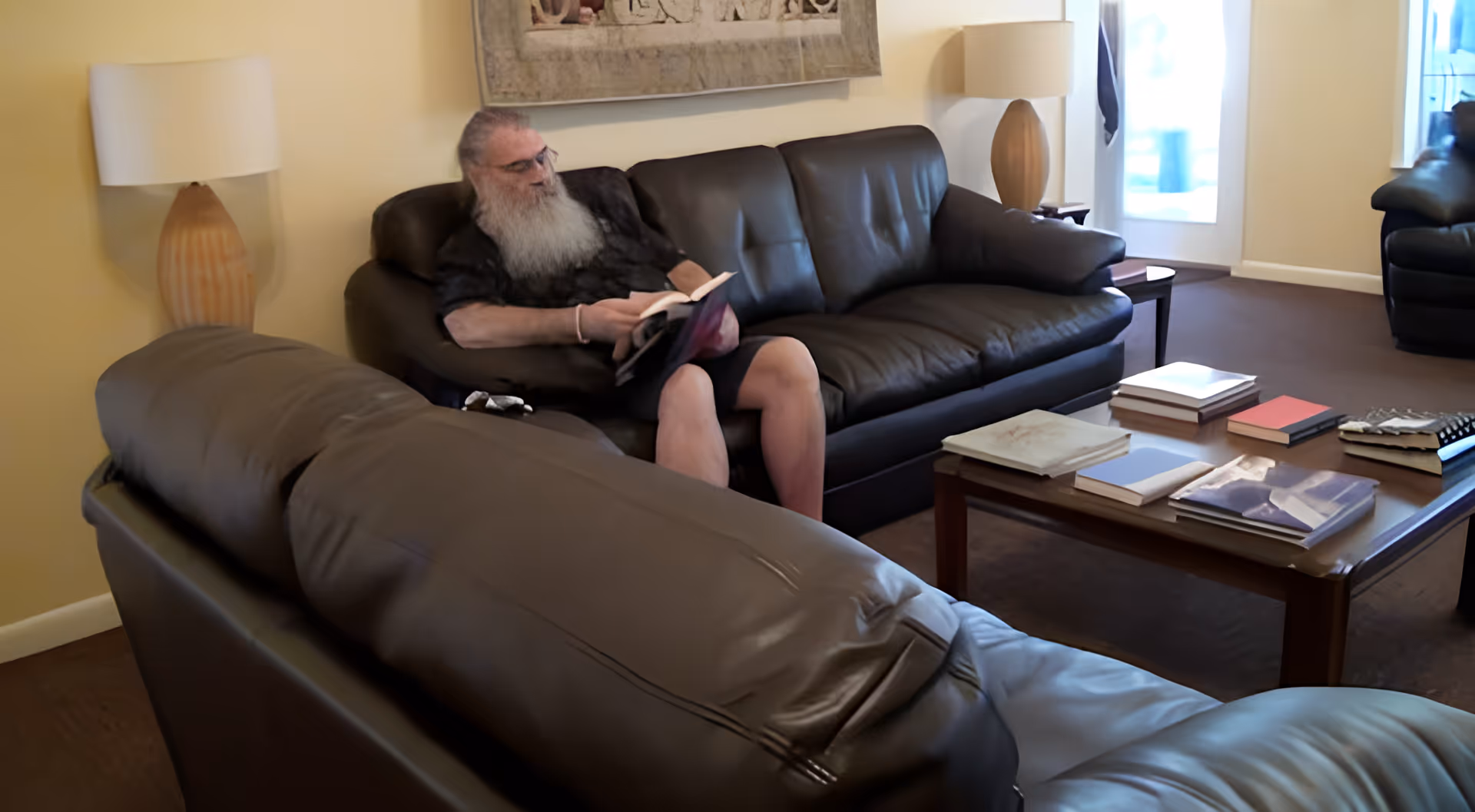 An elderly man with a long white beard is sitting on a black leather couch reading a book in a cozy living room. The room has another black leather couch, a wooden coffee table with several books on it, two table lamps with beige shades, and a large framed artwork on the wall. There is a door with glass panels letting in natural light.