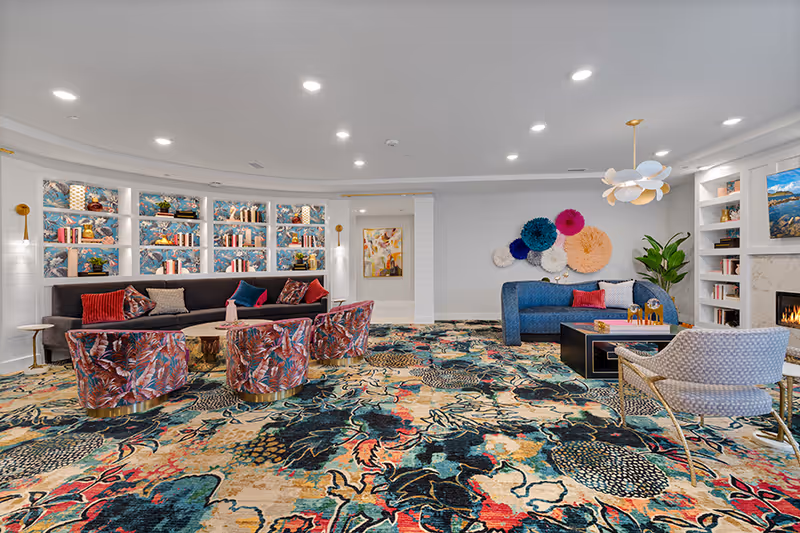 A brightly lit living room with colorful patterned carpet, a dark gray sofa with red and blue pillows, four floral patterned chairs around a wooden coffee table, a blue curved sofa with decorative pillows, a modern white and gold pendant light, a fireplace with a TV above it, built-in shelves with books and decor, and a wall adorned with colorful circular art pieces.
