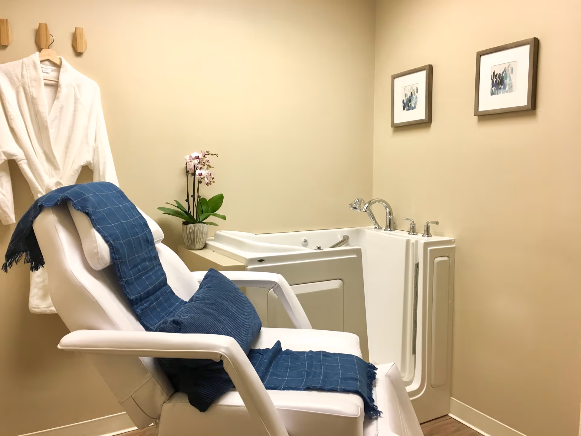 A small spa room with a white reclining chair draped with a blue throw and matching pillow. A white bathrobe hangs on wooden hooks on the wall. There is a walk-in bathtub with chrome fixtures, a potted orchid plant on the ledge, and two framed abstract art pieces on the beige walls.