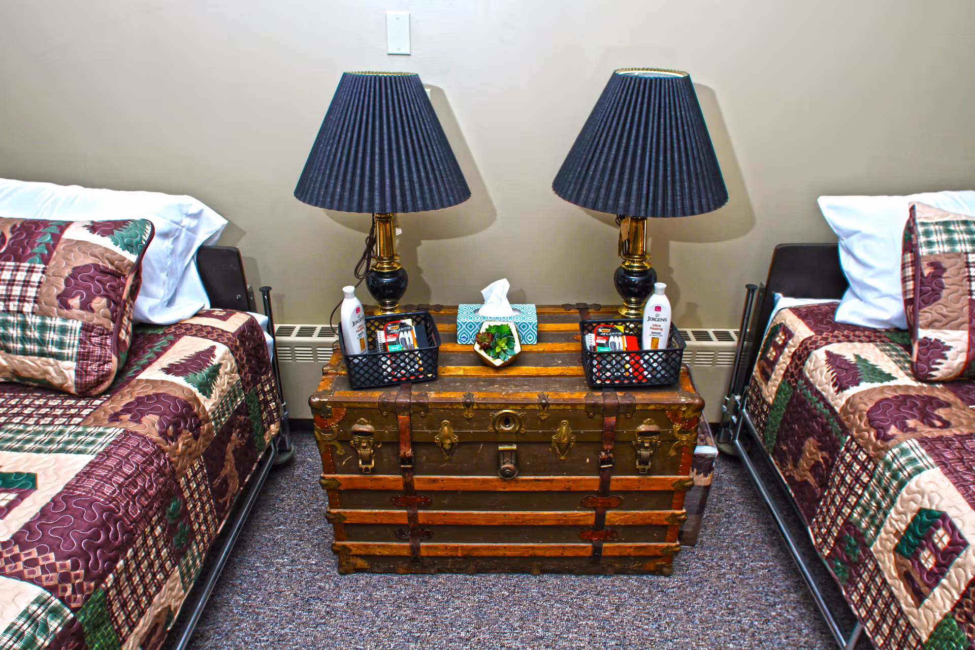 Two twin beds with patterned quilts flank a vintage trunk used as a nightstand topped with two lamps and personal items.