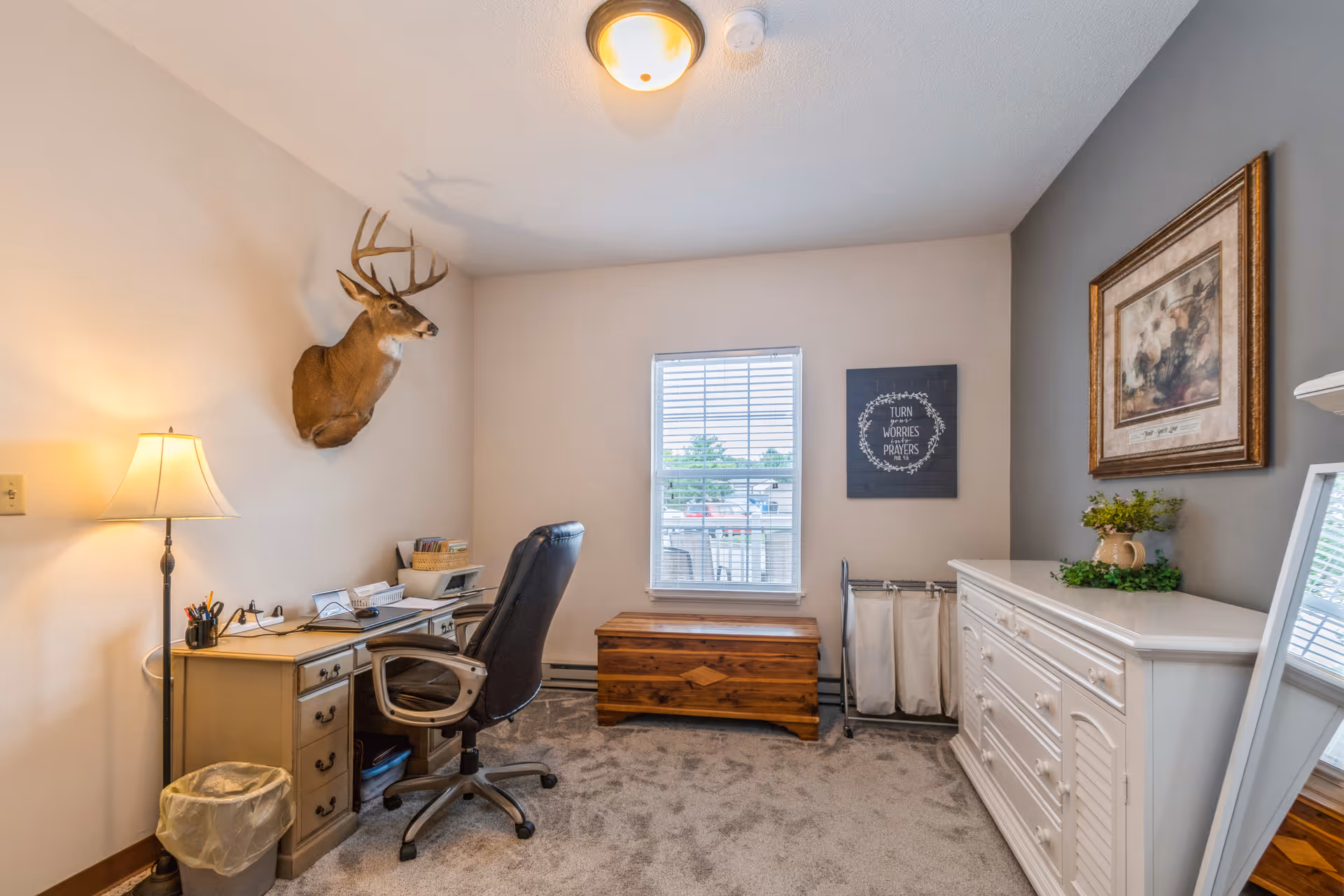 A small furnished bedroom with a desk and office chair, dresser, wooden chest, mounted deer head, and a window.