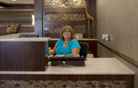 A woman with glasses and a turquoise shirt sitting behind a reception desk in a healthcare facility. The desk has a granite countertop and dark wood paneling with decorative tiles. Behind her is a wall with a decorative giraffe sculpture and a framed photo.
