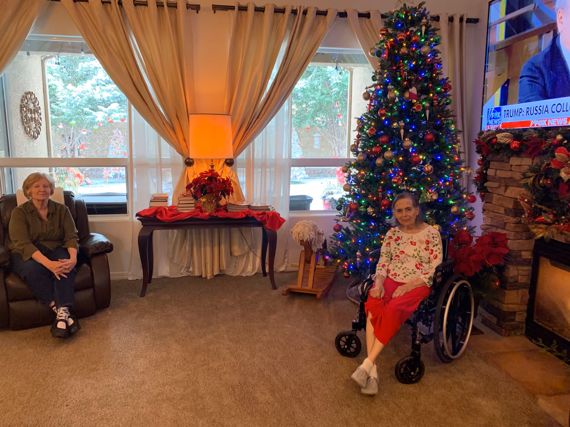 A holiday-decorated living room with two seated people, a large decorated Christmas tree, a lamp-lit table by the window and a TV over a stone fireplace.