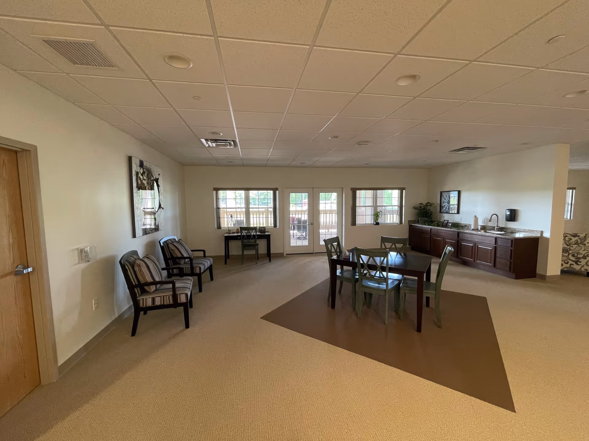A spacious common area with beige carpet and white walls featuring a table with four chairs on a brown rug, two striped armchairs against the left wall, a small desk with a chair near windows and glass doors leading outside, and a countertop with cabinets and a sink on the right side.