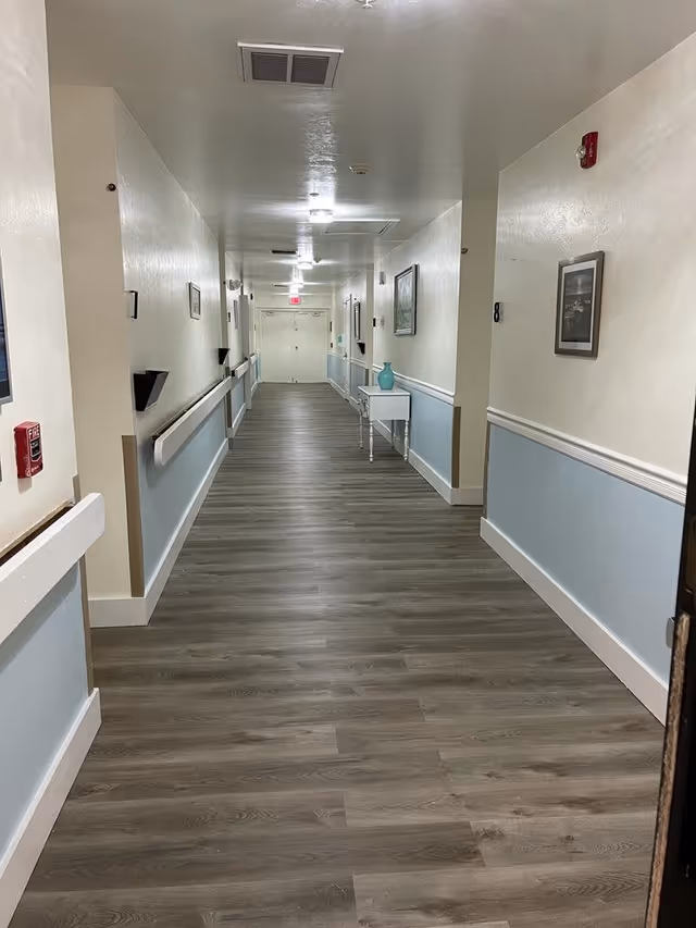 A long, clean hallway in a senior living facility with light blue and white walls, wood-patterned flooring, handrails on both sides, framed pictures on the walls, and a small white table with a blue vase near the end.