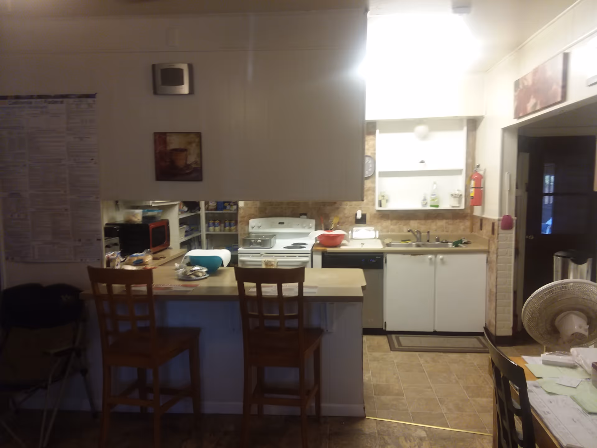 Interior view of a kitchen with a breakfast bar and two wooden stools, a stove and sink, and various items on the countertops.
