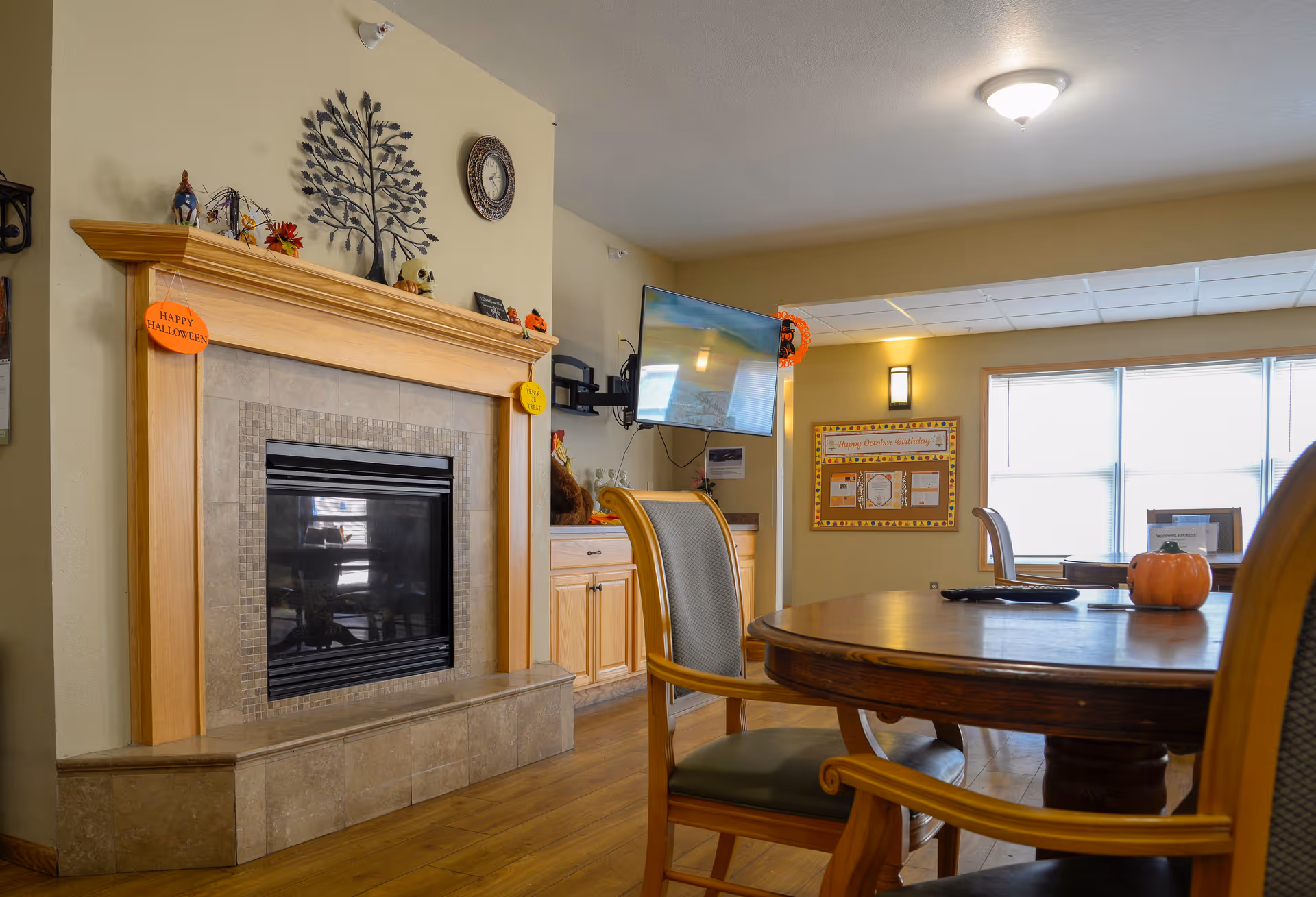 A cozy common area with a wooden round table and chairs, a fireplace with Halloween decorations, a wall-mounted TV, a bulletin board, and a large window letting in natural light.