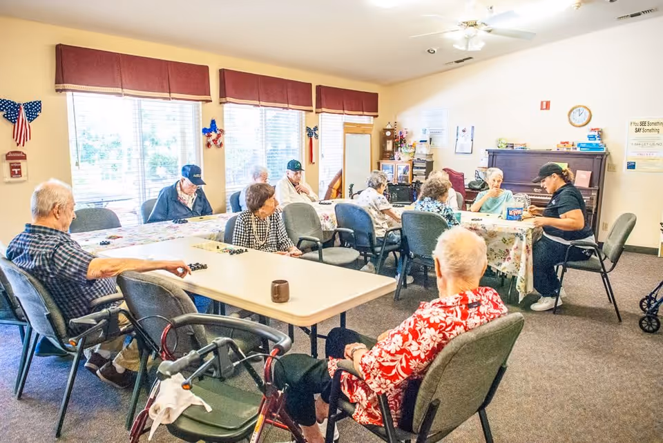 A group of elderly people sitting around tables in a well-lit room with large windows, engaging in activities and conversation. The room has a piano, a clock on the wall, and patriotic decorations.