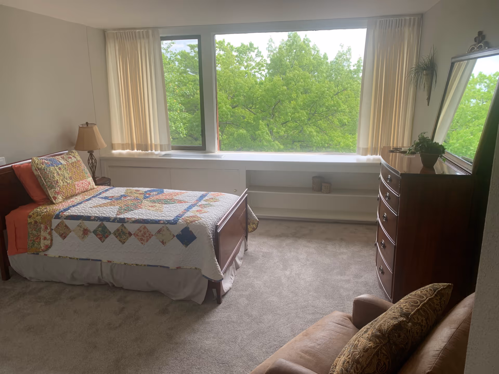 A bedroom with a single bed covered with a colorful quilt and matching pillow. Next to the bed is a small table with a lamp. There is a large window with cream-colored curtains showing green trees outside. A wooden dresser with a mirror and a potted plant is on the right side of the room. In the foreground, there is a cushioned chair with a patterned pillow. The floor is carpeted in a neutral color.