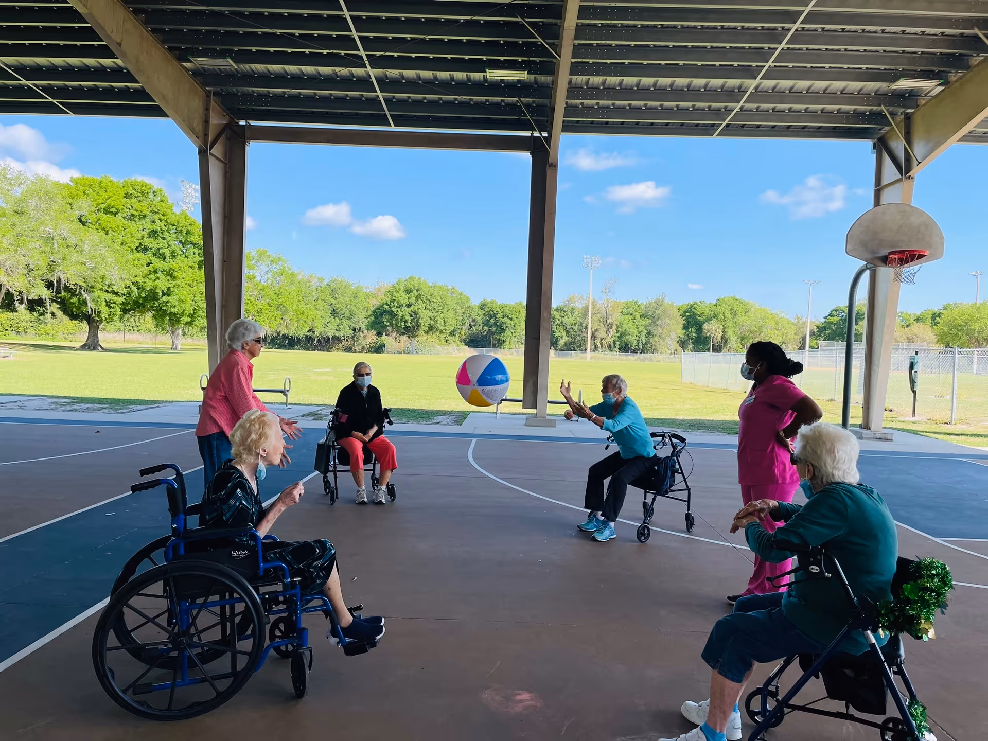 A group of elderly residents and caregivers play with a large beach ball on a covered outdoor basketball court, some seated in wheelchairs or using walkers.