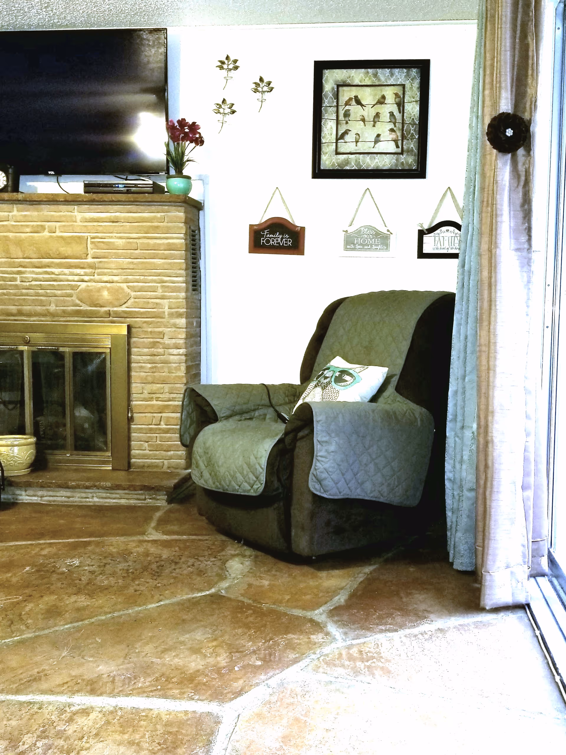 A cozy living room corner featuring a dark armchair with a quilted cover and an owl-themed pillow. To the left is a brick fireplace with a brass-framed glass door, topped by a flat-screen TV and a small green vase with red flowers. The white wall behind the chair has decorative wall hangings including a framed picture of birds and three small plaques with inspirational quotes. Beige curtains partially cover a sliding glass door on the right side.
