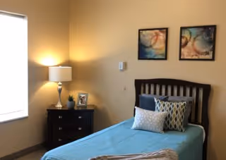 A cozy bedroom with a single bed covered in a blue bedspread and several decorative pillows. Next to the bed is a dark wooden nightstand with a lamp, a small plant, and a framed photo. Two abstract paintings hang on the beige wall above the bed. A window with blinds is on the left side, allowing natural light into the room.