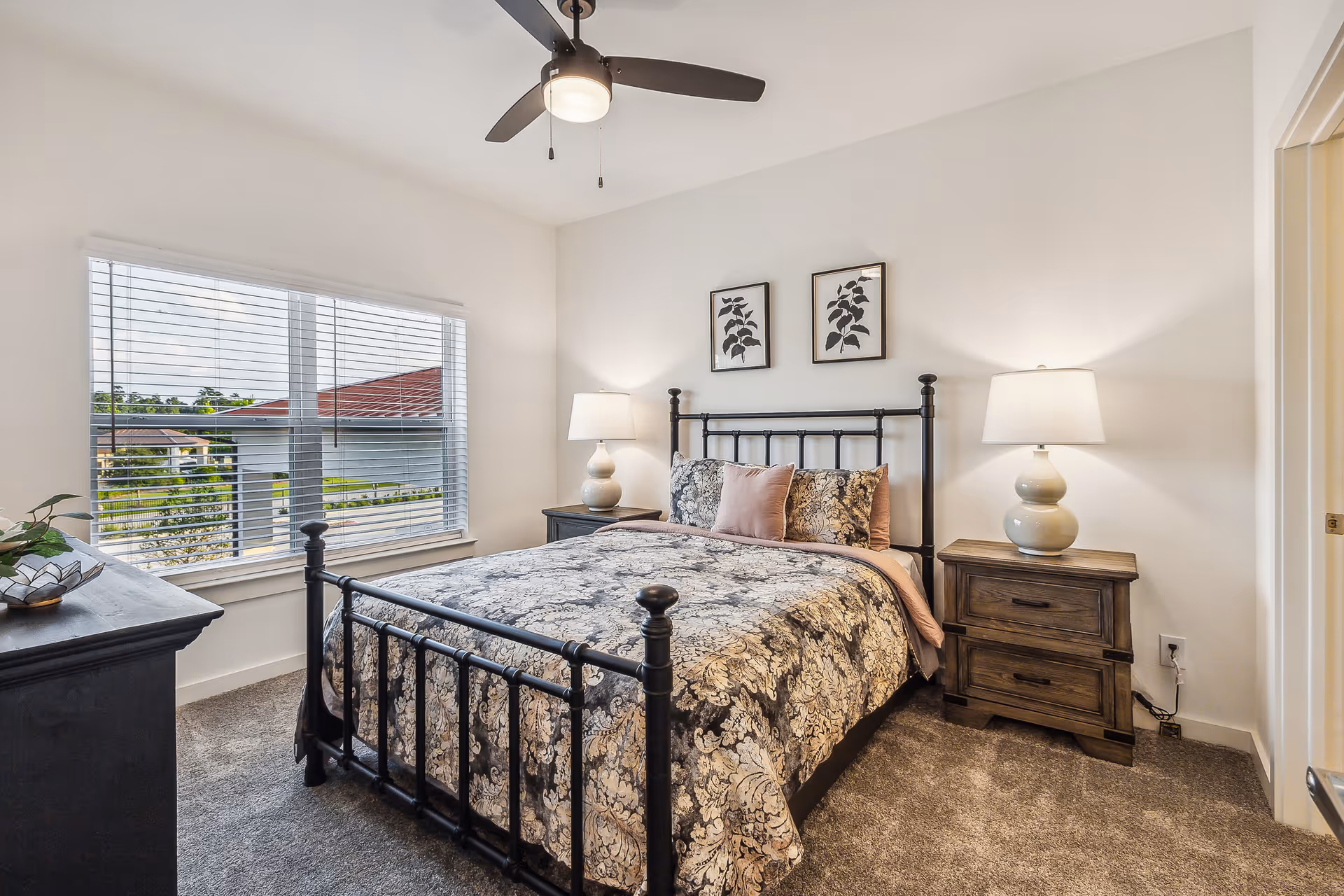 A neatly made bed with a black metal frame and floral patterned bedding in a bright bedroom. Two matching wooden nightstands with white lamps flank the bed. Above the bed are two framed botanical prints. A large window with white blinds lets in natural light, and a ceiling fan with a light fixture hangs overhead. The room has beige carpet and white walls.