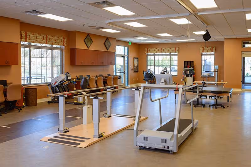 A spacious rehabilitation or physical therapy room with parallel bars for walking exercises, a treadmill, exercise equipment, chairs along the wall, large windows with patterned valances, and warm beige walls with wooden cabinets.