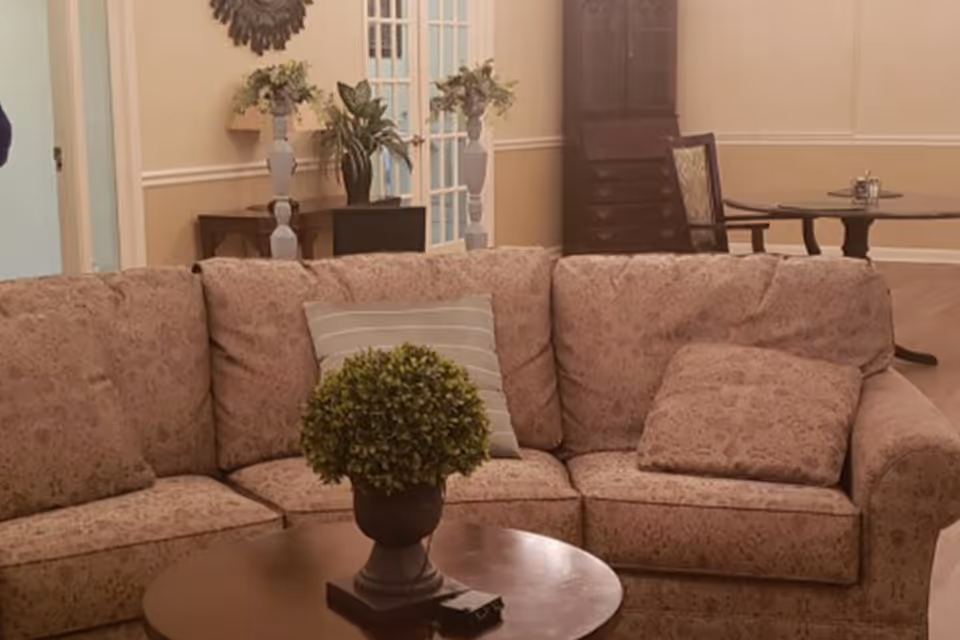 A cozy living room with a patterned beige sectional sofa adorned with two cushions, one striped and one matching the sofa fabric. In front of the sofa is a round wooden coffee table with a small potted plant and a remote control. In the background, there are two tall white vases with flowers on a wooden table, a tall dark wooden cabinet, and a round dining table with chairs. The walls are painted beige with white trim, and there is a decorative wall hanging above the vases.