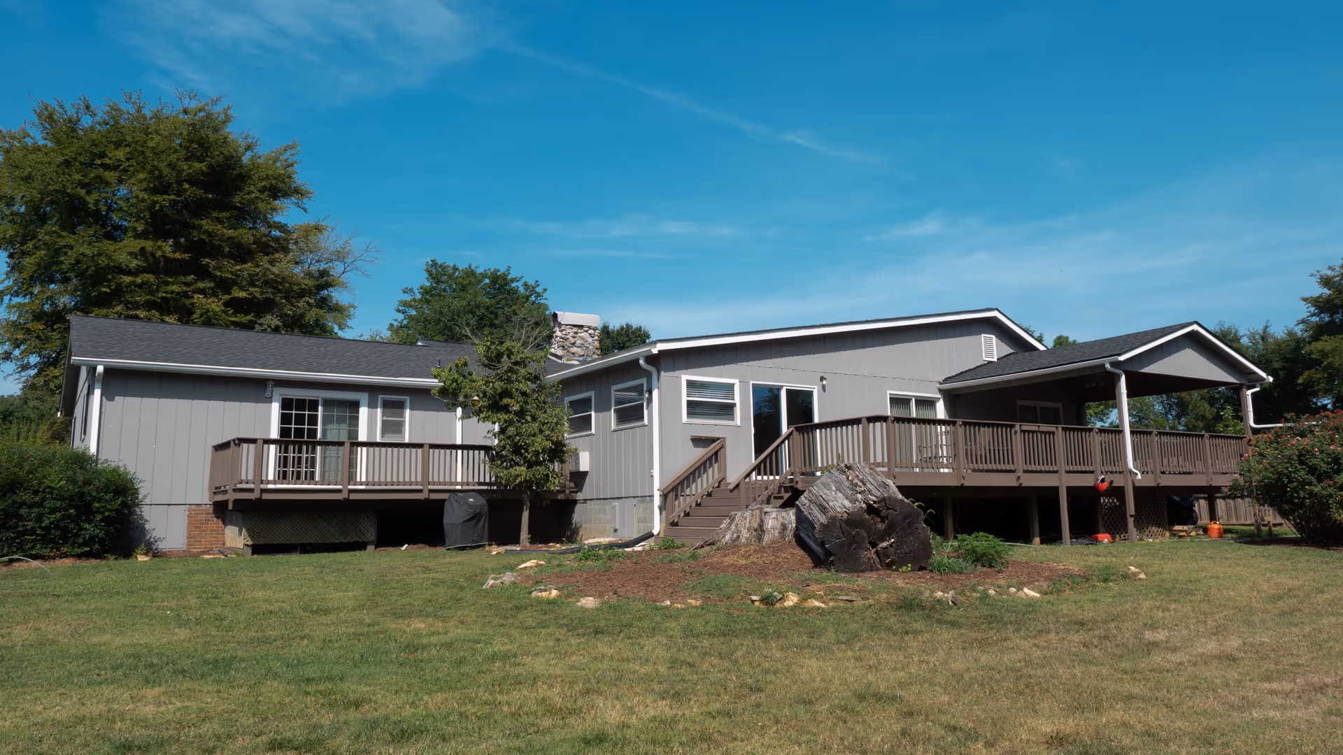 Single-story gray house with a wide wooden deck and steps overlooking a grassy yard under a blue sky.