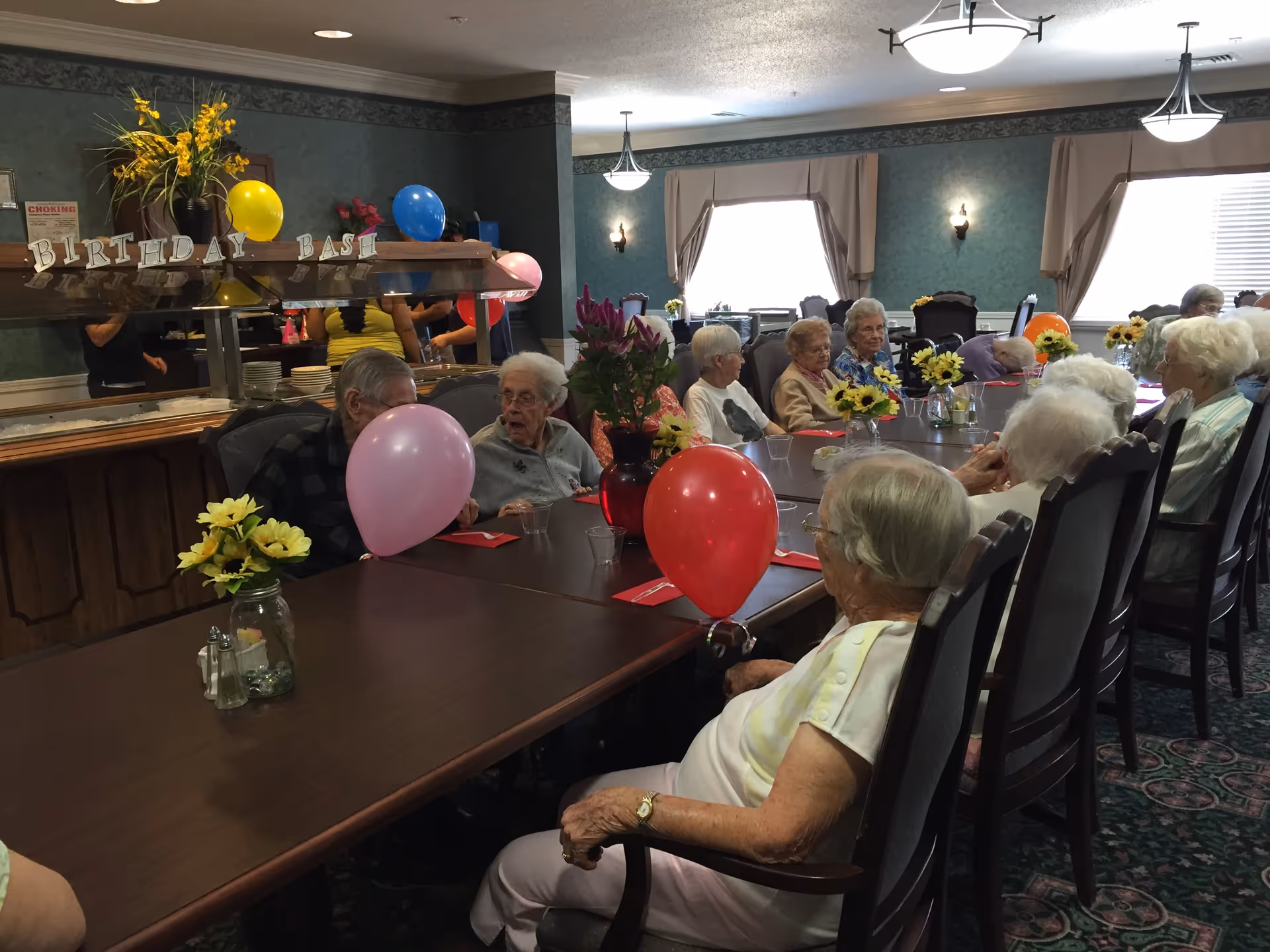 A group of elderly people sitting around a long table in a decorated room celebrating a birthday party with balloons and flowers on the table. A banner reading 'BIRTHDAY BASH' is visible in the background.