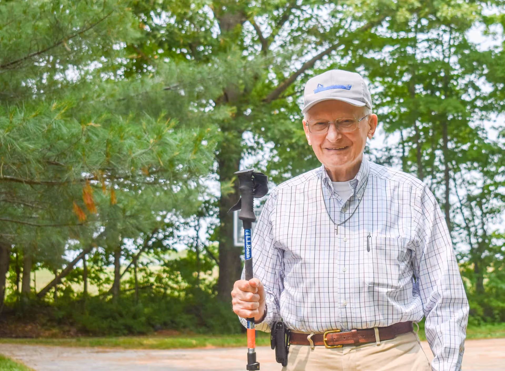 An elderly man wearing a light-colored plaid shirt, beige pants, and a light gray cap is standing outdoors in a wooded area. He is smiling and holding a walking stick. Trees and greenery are visible in the background.