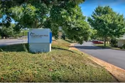 Outdoor view of the entrance area to Meadow Creek Village Assisted Living, showing a sign with the facility's name surrounded by greenery and trees along a paved driveway.