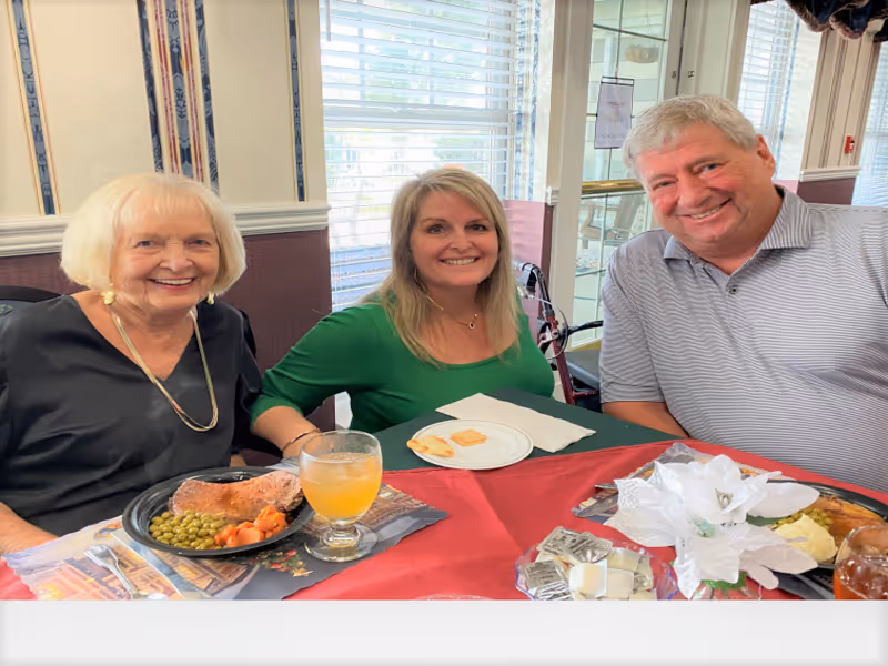 Three people sitting at a dining table in a well-lit room with large windows. An elderly woman on the left is smiling, wearing a black top and gold necklace. A middle-aged woman in the center is wearing a green top and smiling. A middle-aged man on the right is wearing a light gray striped polo shirt and smiling. The table has plates with food, a glass of orange beverage, and a decorative white flower centerpiece.