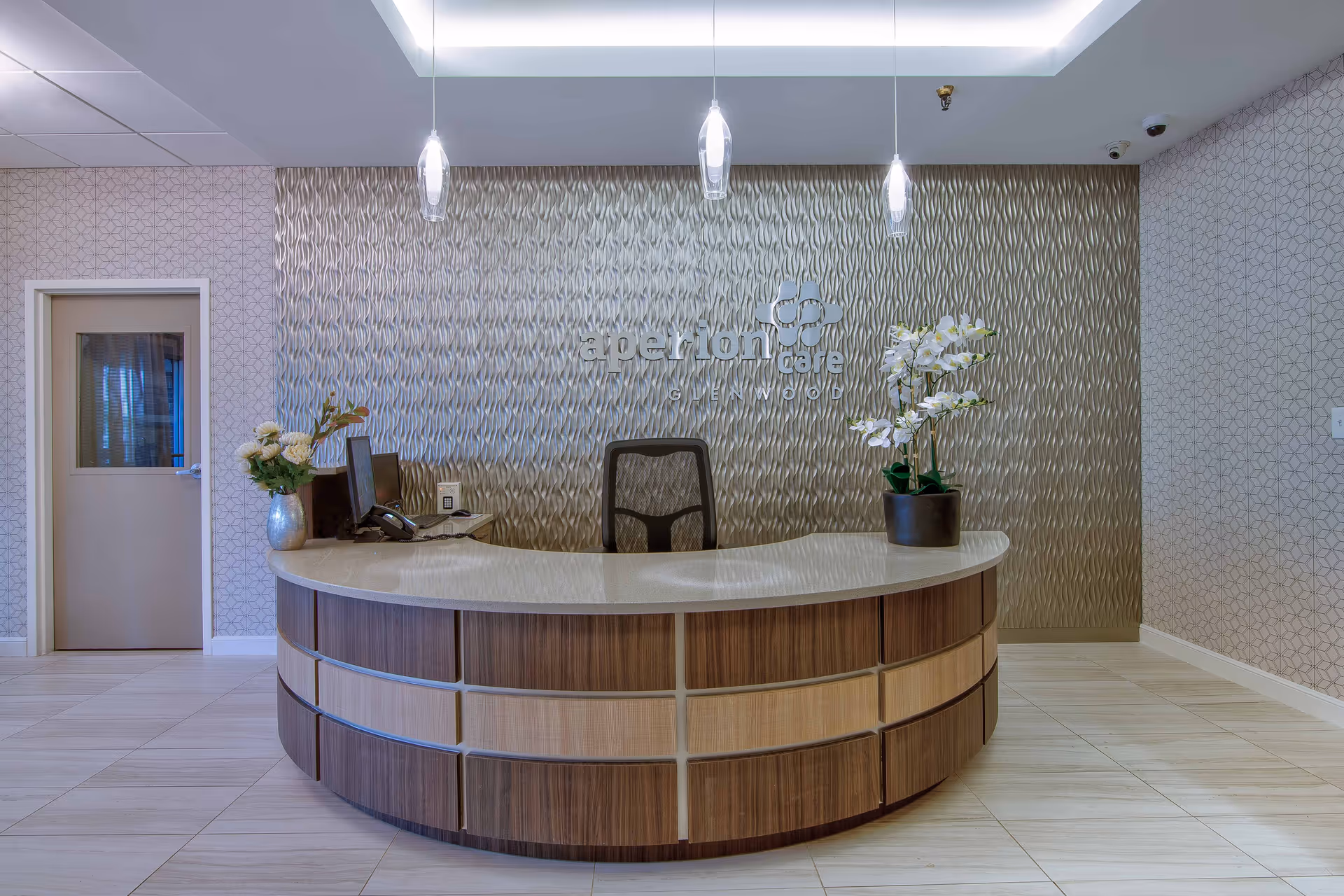 A modern reception desk area with a curved wooden counter and a white countertop. Behind the desk is a textured wall with the sign 'aperion care GLENWOOD'. The desk has a computer, a phone, and two vases with white flowers. Three pendant lights hang from the ceiling above the desk. There is a door to the left and patterned wallpaper on the walls.