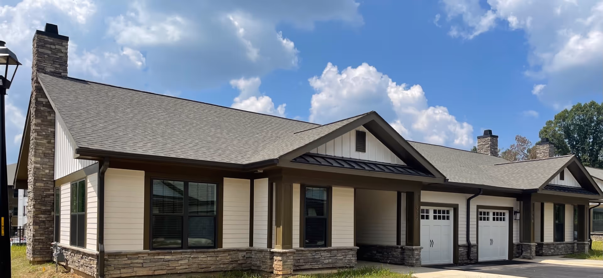 Exterior view of a single-story residential building with beige siding, stone accents, and a gray shingled roof under a partly cloudy blue sky. The building features multiple windows, two white garage doors, and stone chimneys.