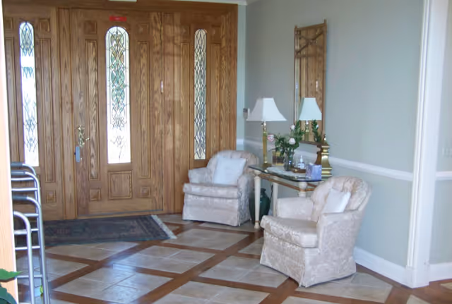 Entryway with wooden double doors, two upholstered armchairs beside a console table holding lamps and flowers, and a tile-and-wood patterned floor.