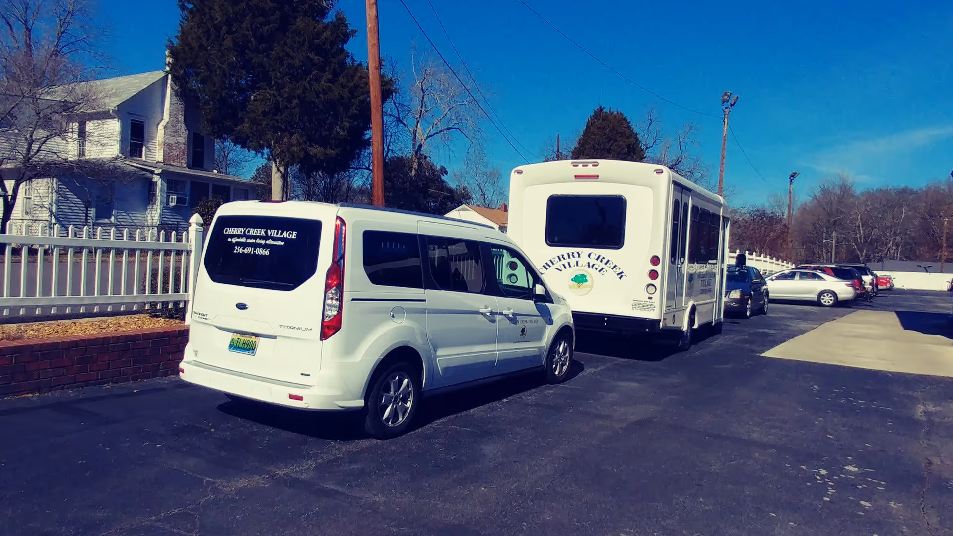 Parking lot with a white van and a white shuttle bus, both labeled Cherry Creek Village, an affordable independent living community for seniors. Several other cars are parked along the lot, with trees and a white fence in the background under a clear blue sky.