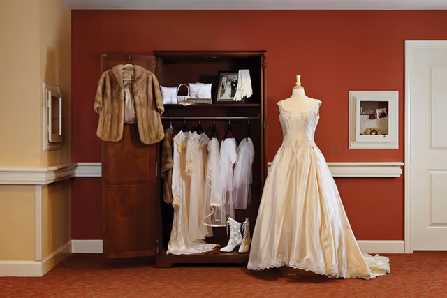 A wooden wardrobe with open doors displaying vintage white dresses, veils, gloves, and accessories. Next to the wardrobe is a mannequin dressed in a white wedding gown. The background features a red wall with a framed display and a closed white door.