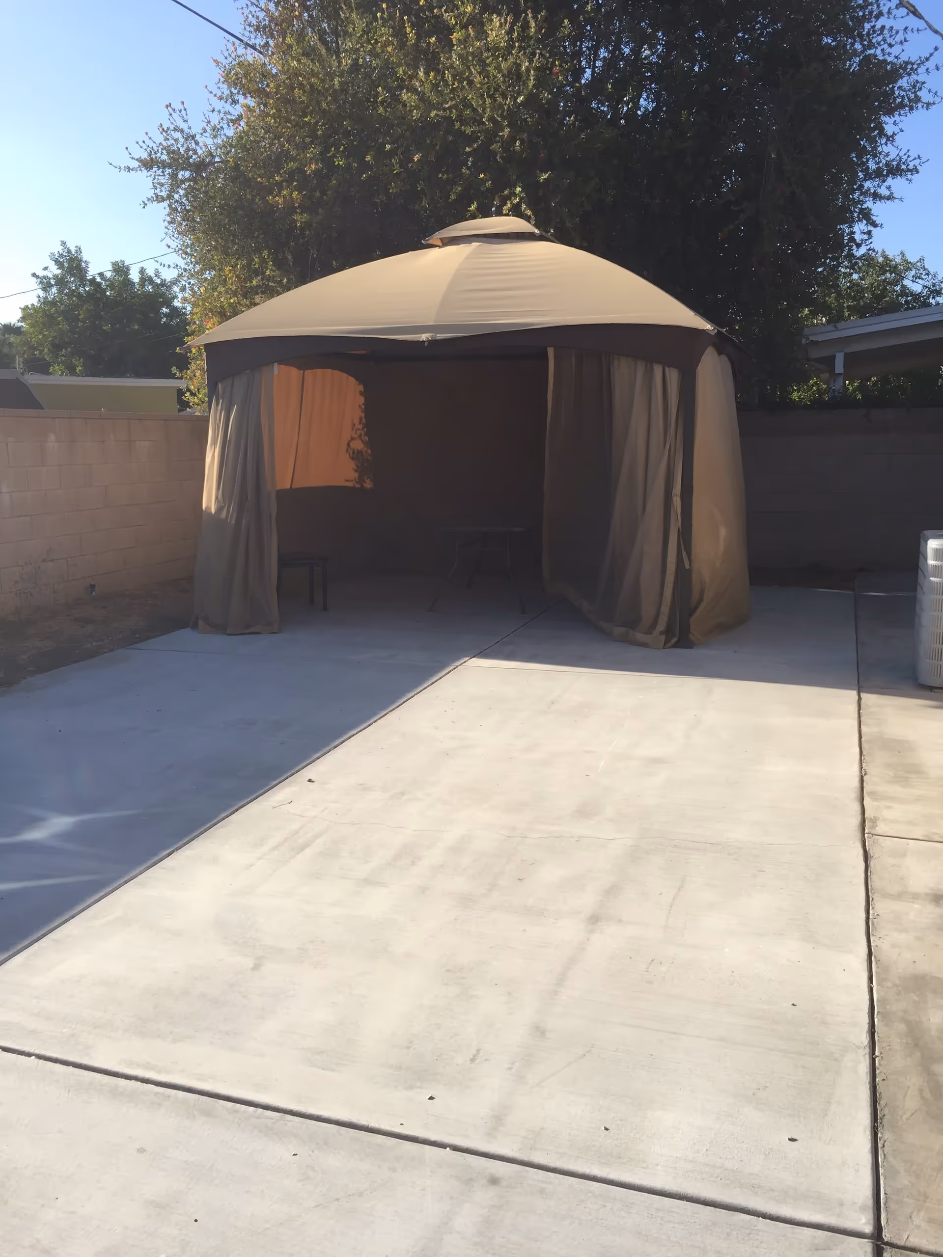 Outdoor patio area with a beige canopy tent set up on a concrete surface. The tent has curtains partially drawn and contains a small table and chair inside. There is a brick wall and trees in the background under a clear blue sky.