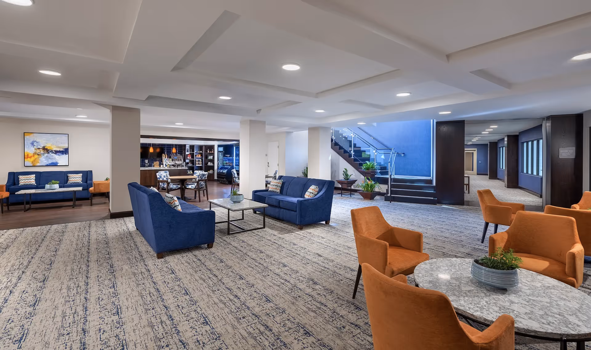 Bright communal lounge featuring blue sofas, orange armchairs, marble-top tables, and a staircase leading to an upper level.