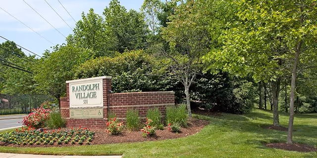 A landscaped outdoor area featuring a brick sign that reads 'Randolph Village 531' surrounded by flowers, shrubs, and trees next to a road.