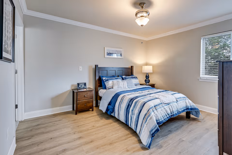 A neatly made bed with blue and white striped bedding in a bedroom with light-colored walls and wood flooring. There is a wooden nightstand with a digital clock and a lamp on it next to the bed. A window with white blinds is on the right wall, and a ceiling fan with a light fixture is above the bed. A framed picture hangs on the wall above the bed.