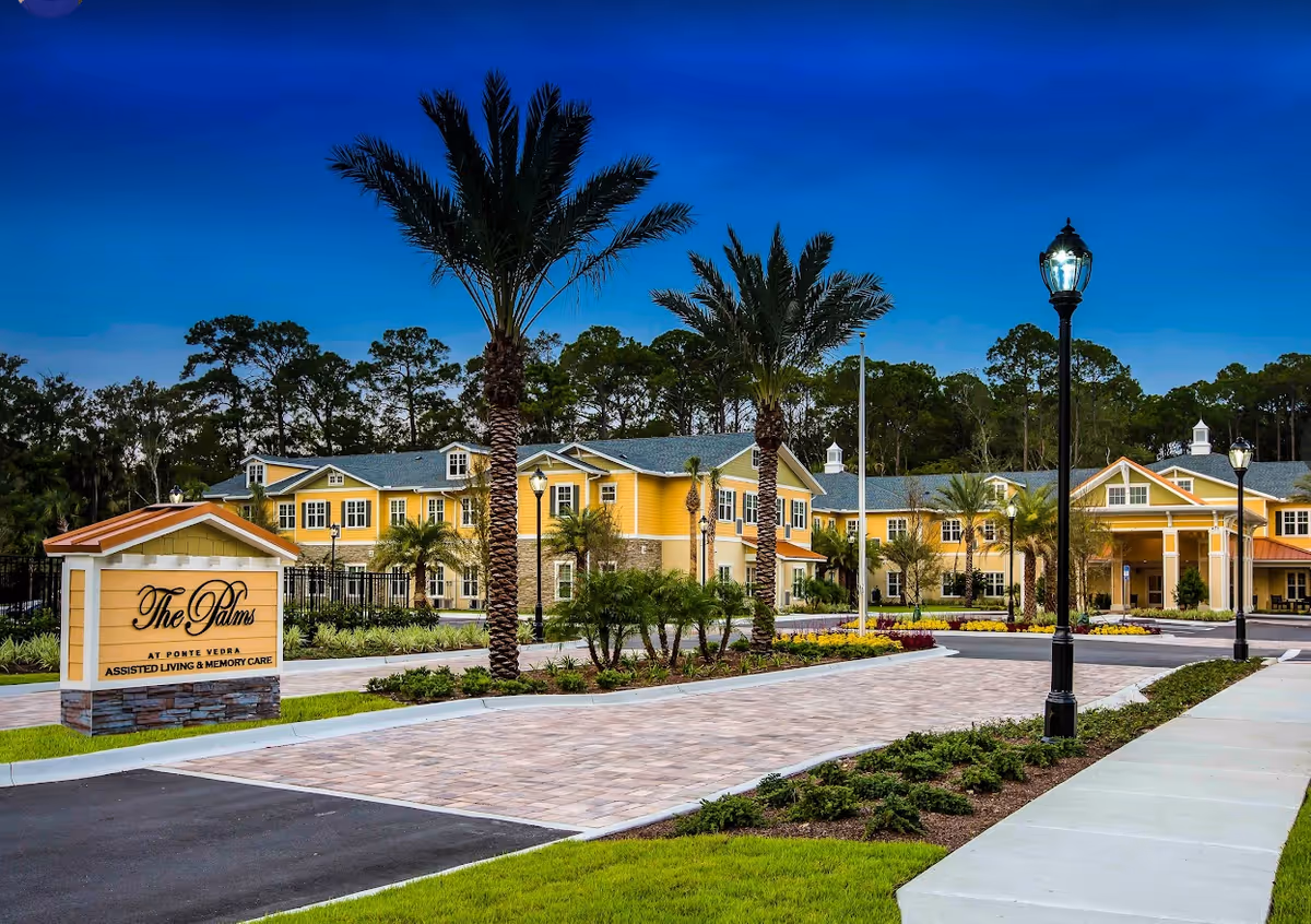 Yellow two-story senior living building front with palm trees, paved driveway, lamp posts, and a sign reading "The Palms."
