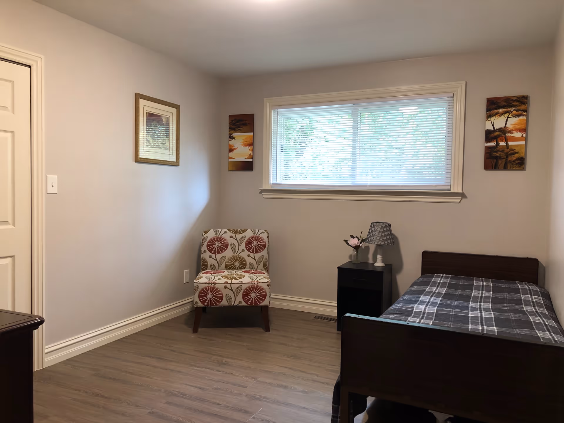 A simple bedroom with a single bed covered with a gray plaid blanket, a small black nightstand with a lamp and a flower vase, a patterned armchair, three framed pictures on the walls, and a window with blinds letting in natural light.
