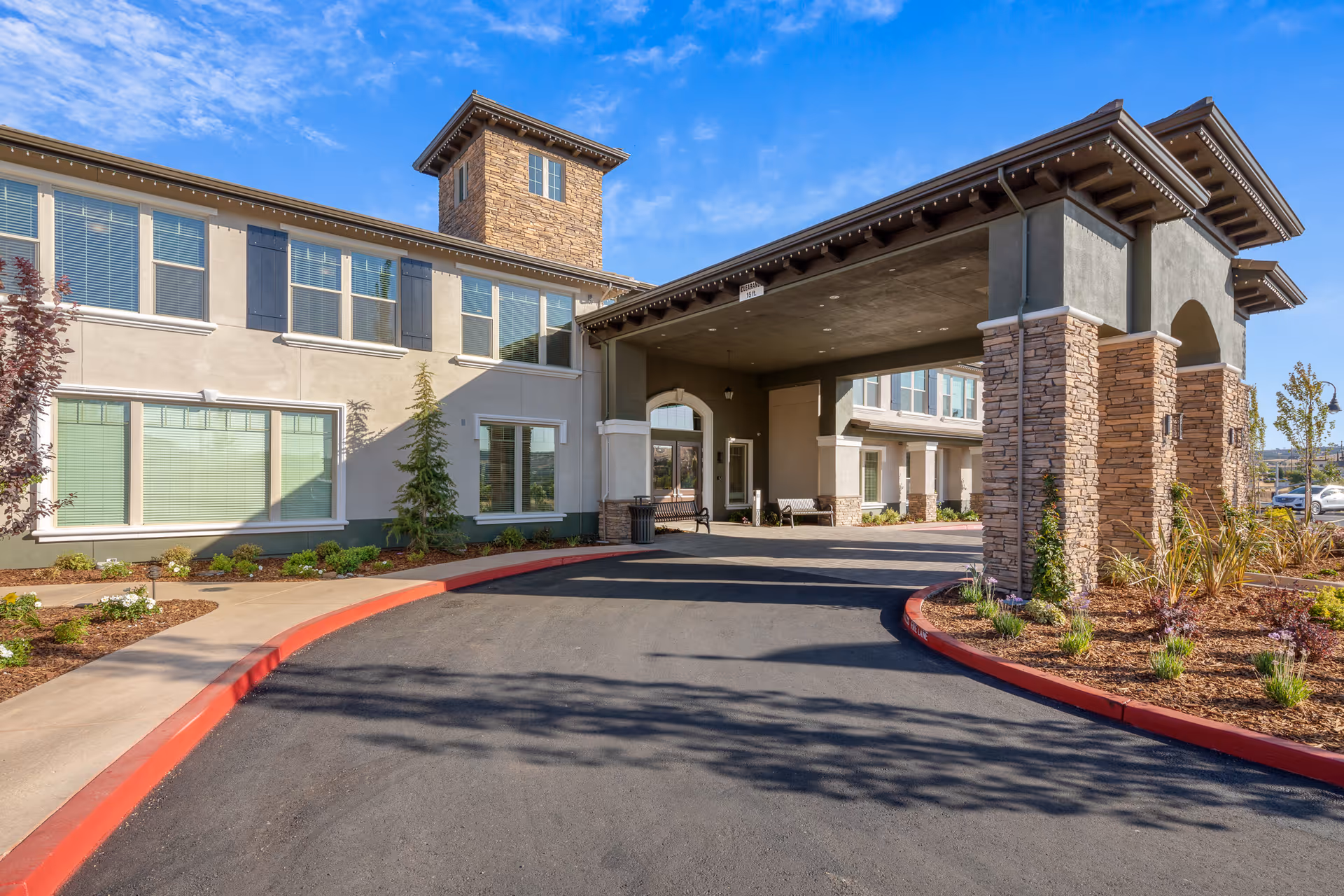 Entrance and porte-cochere of a two-story senior living building with a curved driveway and landscaped beds under a blue sky.